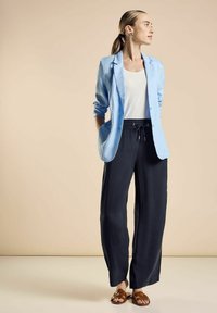 Ljusblå blazer, vit topp, löst sittande mörka byxor med elastisk midja, bruna sandaler. Slät tyg, avslappnad passform, inga mönster eller detaljer.