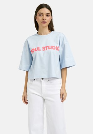 Frau trägt hellblaues, übergroßes T-Shirt mit pinkem "SOUL STUDIO"-Schriftzug und weiße, hoch taillierte Hose, steht vor weißem Hintergrund.