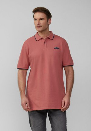 Man in een rozenkleurig poloshirt met marineblauwe accenten en een klein automerk-embleem op de borst, gecombineerd met donkergrijze jeans, staande tegen een effen achtergrond.