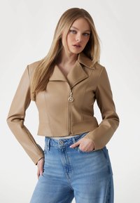 Giacca in finta pelle beige tagliata, con colletto a punte e chiusura con zip. Presenta un accento a logo rotondo e una texture liscia. Abbinata a jeans blu.