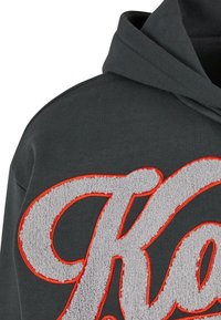Sweat à capuche gris foncé avec de grandes lettres brodées blanches et orange texturées sur la poitrine et une capuche repliée.