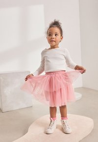 White long-sleeve top, pink tulle skirt, white sneakers with pink accents. Child stands on a textured light surface, playful pose.