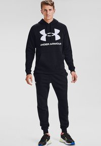 Completo felpa con cappuccio e pantaloni della tuta neri, realizzato in tessuto morbido. Presenta un grande logo bianco di Under Armour sul petto e sui polsini dei pantaloni.