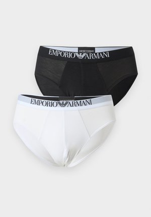 Štíhly tmavokožec muž v bielych slipoch Emporio Armani, stojaci bosý na koberci vedľa kovovej stoličky so sivým vankúšikom, na jednoduchom pozadí.