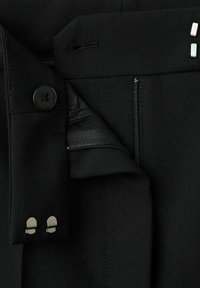 Pantalones de tela negra con una textura suave, que cuentan con un cierre lateral con cremallera, un cierre de botón y acentos metálicos cerca de la cintura.