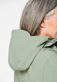 Lichtgroene jas met een capuchon, een zachte textuur en zichtbare stiksels, van opzij getoond met lange grijze haren die gedeeltelijk zichtbaar zijn.