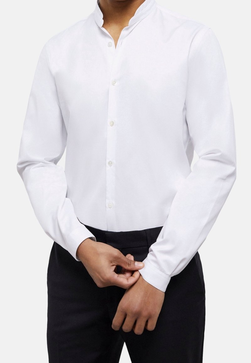 Homme ajustant le poignet d'une chemise blanche ajustée à manches longues, portant un pantalon noir, sur un fond blanc uni.
