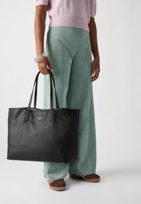 Svart läder totebag med två långa handtag, med en liten guldlogotyp. Modellen har på sig ljusgröna corduroybyxor och bruna skor.