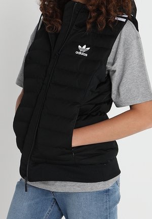 Person trägt eine schwarze gesteppte Adidas-Weste über einem grauen kurzärmligen Shirt und blauen Jeans, die Hände in den Taschen der Weste.