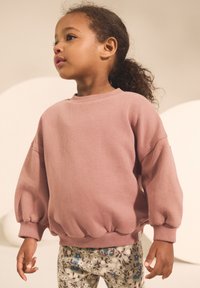 Jong kind met krullend haar in een paardenstaart, gekleed in een losse roze trui en broek met bloemenpatroon, kijkt naar links tegen een neutrale achtergrond.