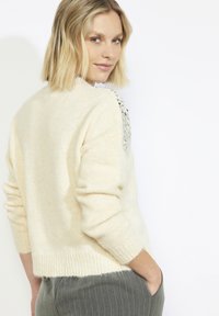 Kreemjas kootud kampsun lahtise lõikega, millel on sädelevate litritega õlaaktsendid ja soonilised mansetid. Pehme tekstuur ja lühendatud ääreosa.