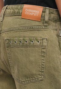 Olívazöld farmernadrág, bőr címkével, rajta "FABIENNE CHAPOT" felirattal. A hátsó zseb fém fűzőjük részletezéssel és zöld fűzős kiegészítőkkel rendelkezik.