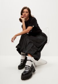 Tricou negru supradimensionat, fustă lungă din satin negru, pantofi cu platformă groasă negri cu trei curele. Scaun transparent, fundal alb.