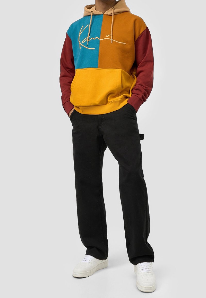 Felpa con cappuccio a blocchi di colore marrone, blu e giallo con cappuccio kaki; presenta una tasca frontale e un logo in corsivo. Abbinata a pantaloni neri e scarpe bianche.