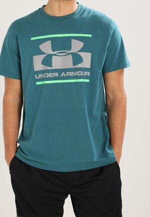 Person iført en teal Under Armour t-shirt med et stort logo og tekst på brystet, kombineret med sorte shorts.