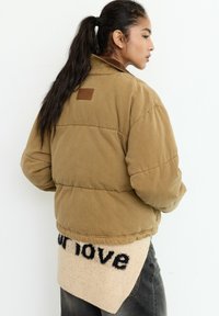 Kvinde med en lang, mørk hestehale iført en brun polstret jakke og en beige sweater med sort tekst, stående sideværts mod en hvid baggrund.