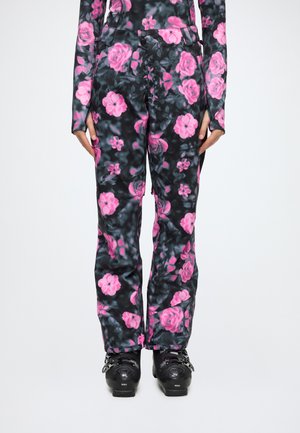 Pantalon style legging avec fond sombre et motif floral rose vif, porté avec des chaussures de ski noires.
