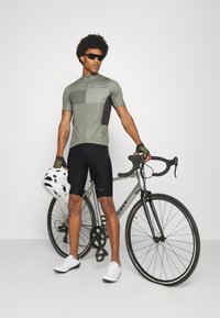 Maillot de cyclisme à manches courtes de couleur olive avec des accents noirs et des rayures horizontales. Cuissard de cyclisme noir, chaussures blanches et casque blanc.