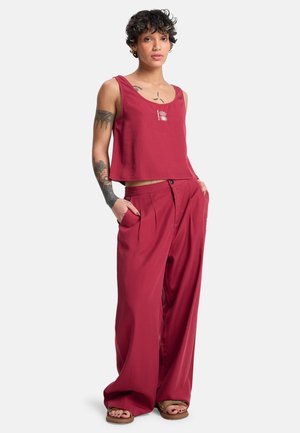 Person mit kurzen lockigen Haaren, Tätowierungen, trägt rotes ärmelloses Oberteil und weite Hosen, steht mit den Händen in den Taschen vor einem einfarbigen Hintergrund.