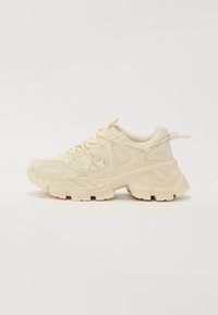 Beige sneaker with mesh accents, thick textured sole, and elastic laces. Features a pull tab at the heel for easy wear.