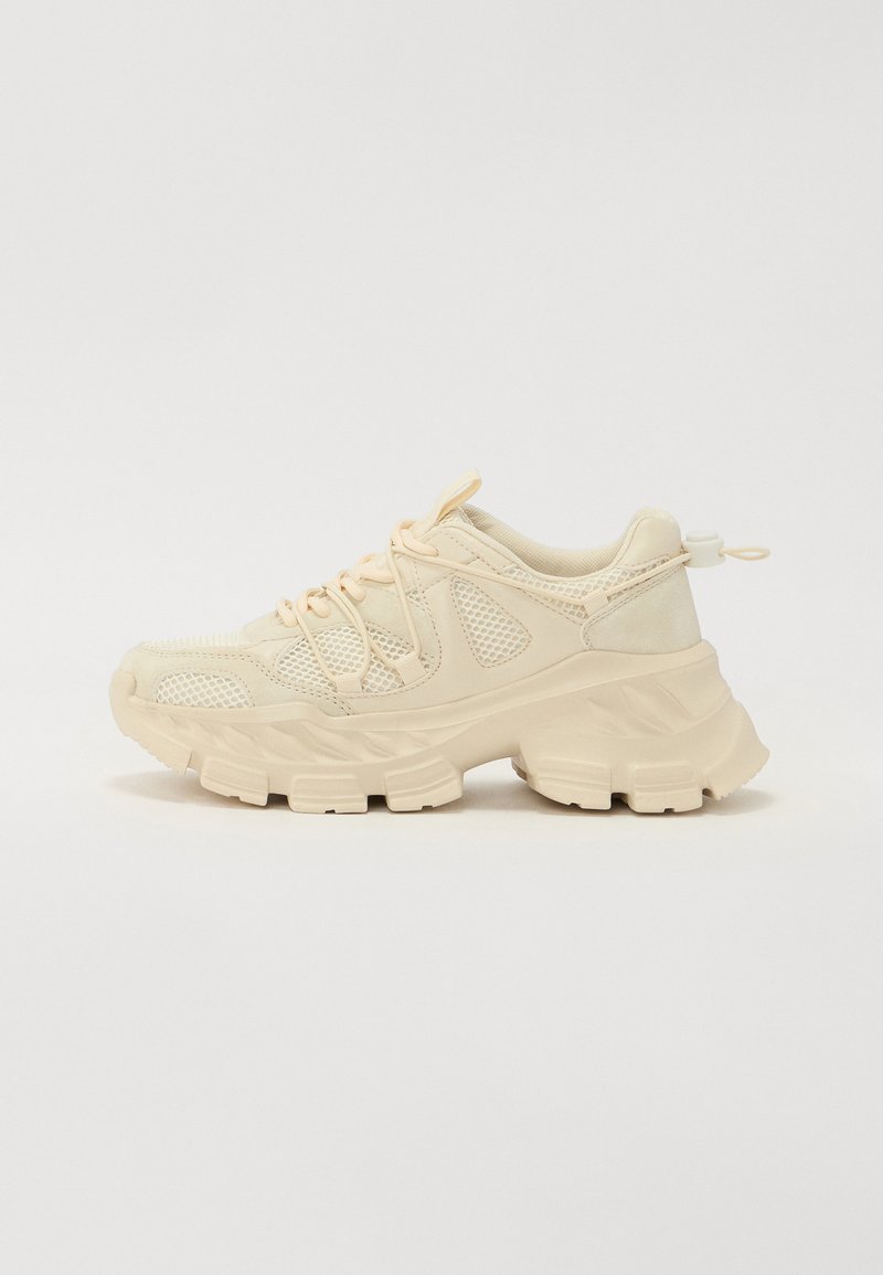 Beige sneaker with mesh accents, thick textured sole, and elastic laces. Features a pull tab at the heel for easy wear.