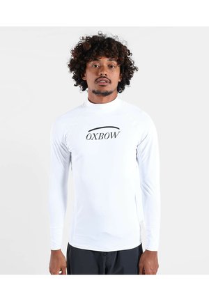 Homme aux cheveux bouclés portant une chemise blanche à manches longues avec le logo "OXBOW" sur la poitrine, debout devant un fond clair uni.