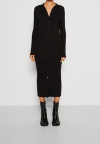 Vestito midi in maglia nera con colletto, maniche lunghe e bottoni sul davanti. Indossato con stivali neri alla caviglia.
