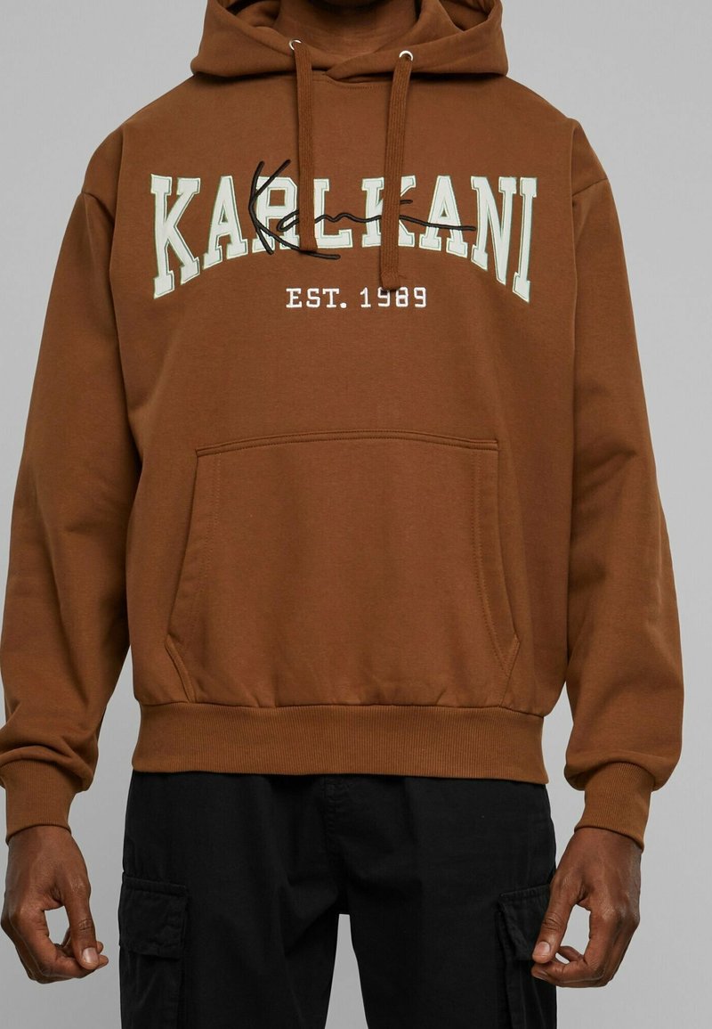 Sweat à capuche marron avec poche kangourou et texte blanc "KARL KANI EST. 1989", porté par une personne à la peau foncée en pantalon noir.