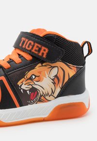 Zapatilla alta negra y naranja con un gráfico de tigre, correa de Velcro, panel de malla texturizada y suela acolchada con un acento naranja.