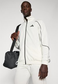 Hvid zip-up atletisk jakke med sorte detaljer, der har høj krave og elastiske manchetter, parret med en sort duffel taske.