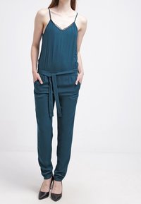 Satin jumpsuit i teal med tunna strappar, spetsdetalj vid halsringningen, bälte i midjan och tapered-byxor. Bärs med svarta klackar. Slät textur.