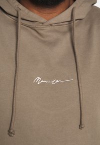 Sudadera marrón con cordones, que presenta una firma bordada en blanco en la parte frontal. Tejido suave con una textura lisa y un corte relajado.