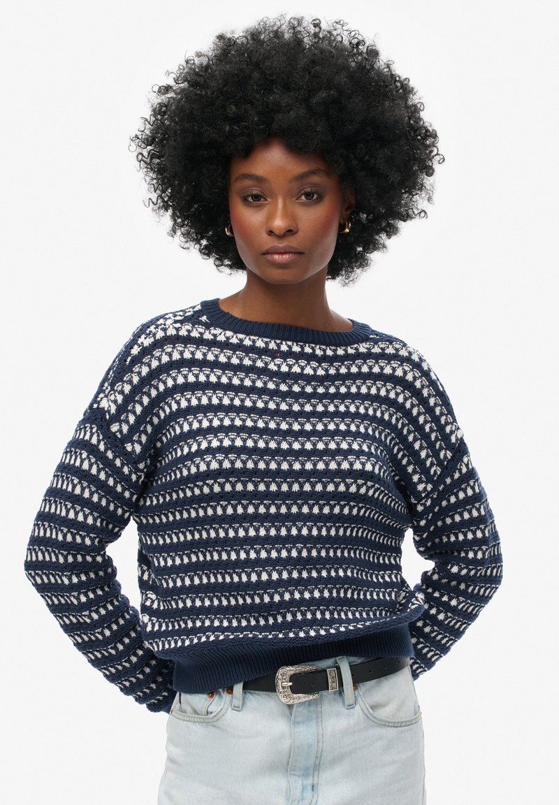 Suéter de punto azul marino y blanco con un patrón de rayas horizontales, cuello redondo y detalle de dobladillo acanalado, combinado con jeans de mezclilla claros.