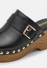 Sabot en cuir noir avec boucle dorée et détails cloutés, doté d'un talon plateforme en bois et d'un dos ouvert.