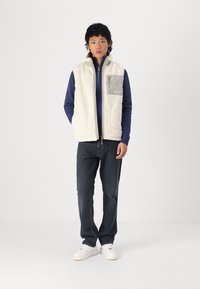 Fleece vest in off-white with gray chest pocket, zipper closure, and side pockets, layered over a blue long-sleeve shirt and dark jeans.