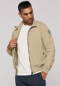 Light beige bomber jacket with a zip front, ribbed cuffs, and a patch on the left sleeve. Smooth fabric texture, styled over a white shirt.