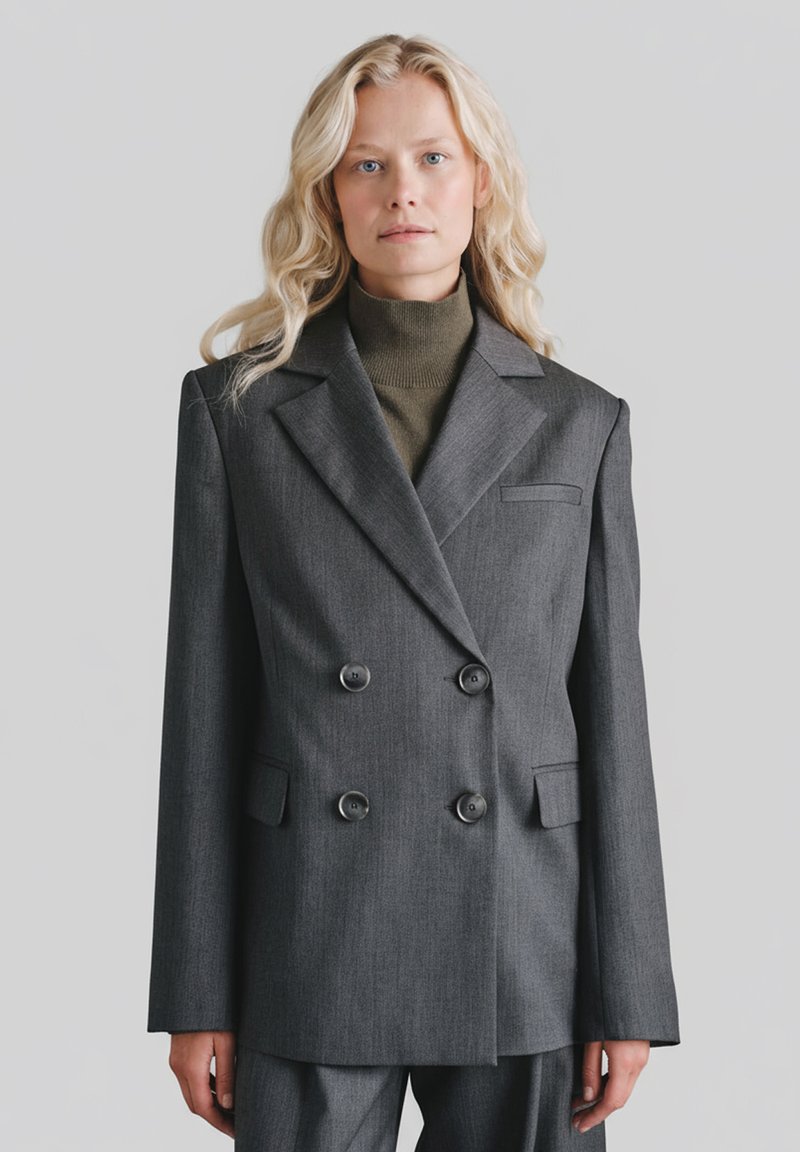 Szary dwurzędowy marynarka z dwoma guzikami, klapami z wycięciem oraz kieszenią na piersi. Noszony na ciemnozielonym golfie. Gładka tekstura tkaniny.