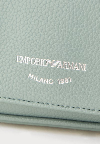Λογότυπο Emporio Armani και έμβλημα αετού εγχαραγμένα σε ασημί σε υφή δέρματος ανοιχτού μπλε με τις λέξεις «Μιλάνο 1981» από κάτω.