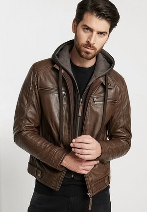 Veste en cuir marron avec une doublure à capuche grise, poches zippées sur la poitrine et design ajusté. Présente des coutures contrastantes et des accessoires en métal argenté.