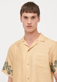 Camisa de lino de manga corta en amarillo claro con bordado en espiral verde en las mangas y un único bolsillo en el pecho.