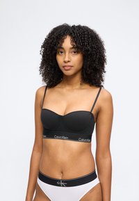 Γυναίκα με σγουρά μαλλιά που φοράει μαύρο σουτιέν Calvin Klein και λευκή εσώρουχα Calvin Klein σε απλό φόντο.