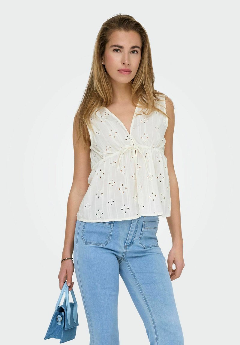 Witte mouwloze top met perforatiepatroon en een gathered strik in de taille, gecombineerd met lichtblauwe jeans en een kleine blauwe handtas.