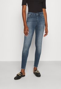 Vaqueros ajustados en denim azul descolorido con un ligero deshilachado en el dobladillo. Combinados con unos zapatos negros de tela con detalles de hardware dorado.