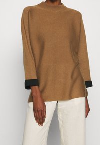 Pull en maille marron avec un col montant et des poignets noirs, offrant une coupe décontractée et une texture côtelée. Associé à un pantalon de couleur crème.