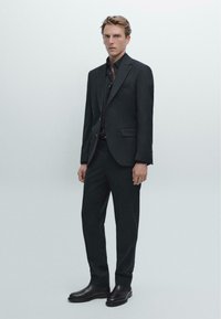 Giacca e pantaloni scuri su misura, abbinati a una camicia nera. Tessuto liscio, vestibilità slim, design minimalista e stivali neri eleganti.