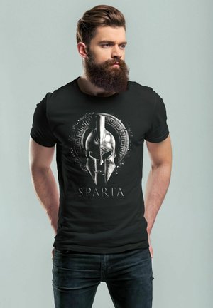 Schwarzes T-Shirt mit einem silbernen Spartaner-Helm-Grafik, umgeben von einem kreisförmigen Rand, verziert mit einem griechischen Muster, und dem Wort "SPARTA" in fetter Schrift.