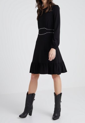 Vrouw die een zwart jurk tot op de knie draagt met gestikte taille details en lange mouwen, gecombineerd met zwarte haklaarzen tot halverwege de kuit.