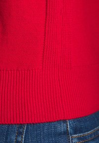 Gros plan sur l'ourlet d'un pull rouge en maille au-dessus d'un jean en denim bleu avec des coutures visibles.