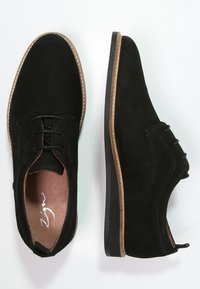 Un paio di scarpe da uomo in suede nero con soletta in pelle marrone e cuciture beige lungo i bordi della suola su uno sfondo bianco.