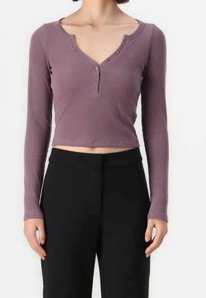 Kobieta ubrana w fioletowy, prążkowany, długi rękaw, krótki top z guzikami oraz czarne spodnie z wysokim stanem, stojąca na tle jednolitego tła.